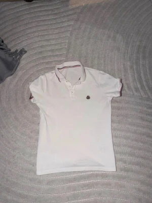 Vit pikétröja från Moncler - Vit pikétröja från Moncler med klassisk krage och knäppning framtill. Tröjan har röda och blå detaljer på krage och ärmslut samt Moncler-logga på bröstet. Perfekt till en clean och sportig stil.