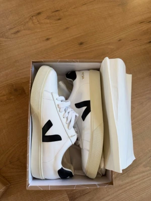 Vita/Svarta Veja Campo 46 - Stilrena vita sneakers från Veja med svart V-logga på sidan och svarta detaljer vid hälen. Skorna har klassisk låg modell, vita skosnören och ovandel i läder. Perfekta för en clean och trendig look. Pris kan diskuteras vid snabb affär.