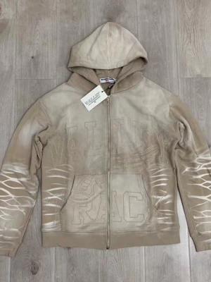 Beige zip hoodie från Valley Track - Snygg beige hoodie från Valley Track med dragkedja framtill och huva. Hoodien har ett coolt mönster med vita detaljer på ärmarna och stor broderad text på bröstet. 