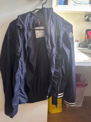 Marinblå vindjacka från Moncler - Snygg marinblå vindjacka från Moncler med dragkedja, ribbade muddar med vita ränder och klassisk Moncler-logga på ärmen. Jackan har en sportig look och är tillverkad i lätt, glansigt material som passar perfekt till streetwear och casual outfits.
