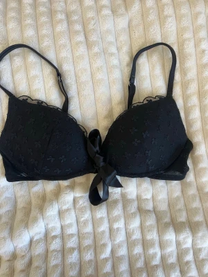 Svart push-up bh med rosett - Snygg svart push-up bh med spetsdetaljer och små blommor i tyget. Dekorerad med en stor satinrosett framtill och justerbara axelband. Perfekt för dig som vill ha en feminin och stilren look.