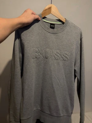Grå sweatshirt från Hugo Boss - Snygg grå sweatshirt från Hugo Boss med broderad logga framtill. Tröjan har rund halsringning, långa ärmar och ribbade muddar. Perfekt för en avslappnad och stilren look. Fråga om du vill ha mer bilder!