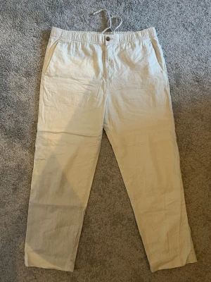 Beiga linnebyxor  - Snygga beiga linnebyxor med avslappnad passform och dragsko i midjan. Byxorna har sidfickor och stängs med knapp. Perfekta för en chill och stilren look under varma dagar. Tillverkade i lätt och luftigt linnematerial.