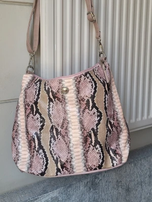 Rosa axelremsväska i ormmönster crossbody rymlig  - Säljer en snygg handväska med rosa och beige ormmönster och justerbar axelrem i ljusrosa. Väskan har en rund form och stängs med tryckknapp. Perfekt för dig som gillar djurmönstrat och vill sticka ut lite extra.
