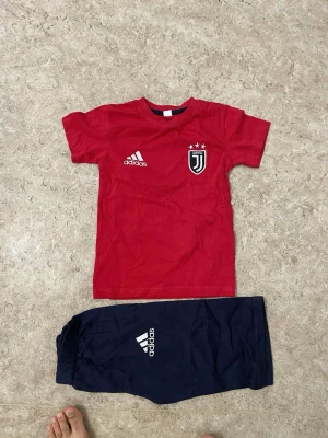 Röd Adidas Juventus t-shirt och blå shorts - Säljer ett set med röd t-shirt från Adidas med Juventus-logga och tre stjärnor på bröstet, samt blå shorts med Adidas-tryck. Perfekt för träning eller sport. Materialet är mjukt och lätt, t-shirten har rund hals och korta ärmar.