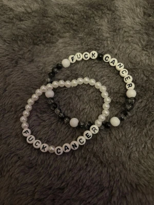 Pärlarmband med text Fuck Cancer 2 Pack - Två elastiska armband med vita och svarta pärlor samt bokstavspärlor som stavar 'FUCK CANCER'. Ena armbandet har vita och svarta pärlor, det andra har vita pärlor. Perfekt statement-accessoar för att visa stöd och styrka.