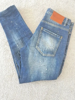 Blå distressed jeans från Dsquared2 - Säljer ett par blå jeans från Dsquared2 med slitningar, hål och vita färgstänk. Jeansen har klassisk femficksdesign, läderpatch bak och en snygg tvättad look. Perfekt för dig som gillar en edgy och trendig stil.