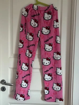 Hello Kitty rosa pyjamasbyxor - Supermjuka och mysiga pyjamasbyxor i rosa fleece med Hello Kitty-motiv och text. Byxorna har resår i midjan och är perfekta för chilliga kvällar hemma. Sött tryck med Hello Kittys ansikte och röda rosetter över hela tyget.