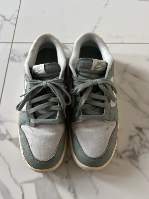 Nike grå låga sneakers - Snygga låga sneakers från Nike i grått med vita detaljer. Ovandelen är en mix av mocka och läder, med klassisk Nike-logga på tungan. Skorna har rund tå och platt sula med tydligt mönster för bra grepp. Perfekta för en avslappnad streetstil.