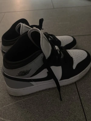 Svarta och vita Air Jordan 1 Mid - Snygga Air Jordan 1 Mid sneakers i svart, vitt och grått läder. Klassisk siluett med perforerad tå, svart swoosh och ikoniska Jordan-loggor på sidan och plösen. Skorna har svarta skosnören och en vit sula som ger en clean look.