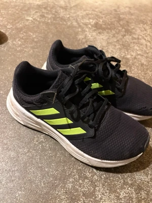 Svarta Adidas sneakers med neonränder - Svarta sneakers från Adidas med klassiska tre neonfärgade ränder på sidorna. Ovandelen är i meshmaterial för bra andningsförmåga och sulan är vit med sportig känsla. Perfekta för en aktiv livsstil och snygga till streetwear.