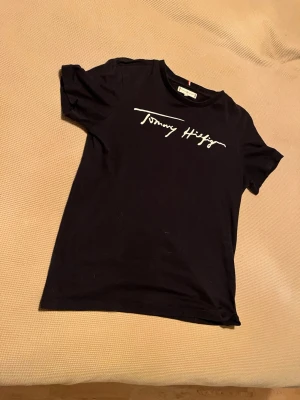 Svart Tommy Hilfiger t-shirt - Snygg svart t-shirt från Tommy Hilfiger med vit logga i skrivstil på bröstet. Klassisk passform och rund halsringning, perfekt till jeans eller shorts. Tillverkad i mjuk bomull för skön känsla hela dagen.