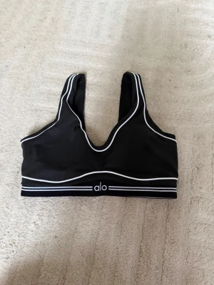 Svart sport-bh från Alo med vita detaljer - Säljer en svart sport-bh från Alo med vita kontrastlinjer och breda axelband. Bh:n har en djup rundad halsringning och elastiskt band nedtill med Alo-loggan. Materialet är mjukt och stretchigt, perfekt för träning eller att ha hemma 🤍