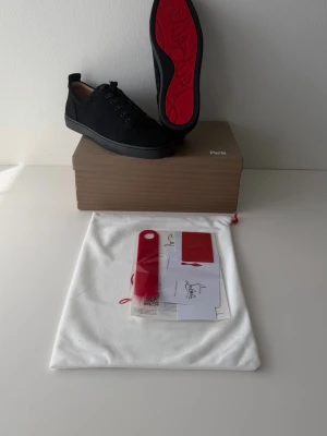 Svarta Christian Louboutin sneakers - Snygga svarta sneakers från Christian Louboutin med klassisk röd sula. Skorna har en låg profil, rund tå och svarta skosnören. Tillverkade i textil och läder, med märkets logga på tillbehören. Perfekta för dig som gillar exklusiva detaljer och streetstyle.