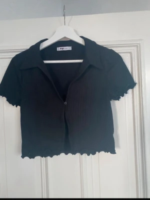 Svart cropped topp från Primark - Svart ribbad cropped topp från Primark med korta ärmar, krage och en knapp framtill. Toppen har vågig kant nertill och vid ärmslut, vilket ger en trendig look. Perfekt att styla med jeans eller kjol.