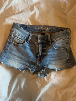 Blå jeansshorts från American Eagle - Snygga blå jeansshorts från American Eagle med råa kanter och klassisk femficksdesign. Shortsen har låg midja och en avslappnad passform, perfekta för varma sommardagar. Ikonisk denimlook med slitna detaljer och knappgylf.