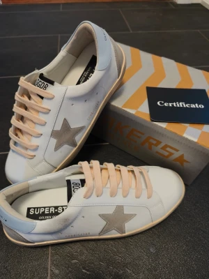 Golden Goose Superstar storlek 42 - NYPRIS ca 6000kr, mitt pris 1699kr. Säljer billigt pga att jag bytte skosnörena med en kompis. Golden Goose Superstar sneakers i vitt läder med beige mockadetaljer och grå stjärna på sidan. Klassisk låg modell med beige sula och vita skosnören. Märkeslogga på plösen och hälpartiet. Snygg och trendig design för dig som gillar streetstyle. 