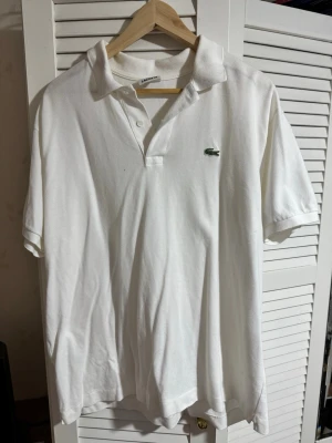 Vit pikétröja från Lacoste - Klassisk vit pikétröja från Lacoste med korta ärmar och krage. Tröjan har knappar framtill och den ikoniska gröna krokodilloggan på bröstet. Perfekt för en stilren och sportig look.