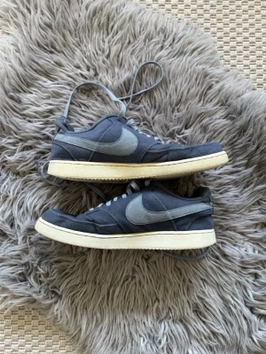 Blå Nike sneakers med vit sula - Snygga låga sneakers från Nike i mörkgrå textil med ljusblå swoosh och vit sula. Klassisk design med snörning och retrovibbar. Perfekta för dig som gillar stilrena och tidlösa sneakers.