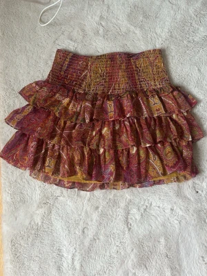 Mönstrad volangkjol från Zara - Färgstark kortkjol från Zara med paisleymönster i rött, gult och lila. Kjolen har flera lager volanger och en smockad midja som ger en snygg och bekväm passform.  Pris kan diskuteras!