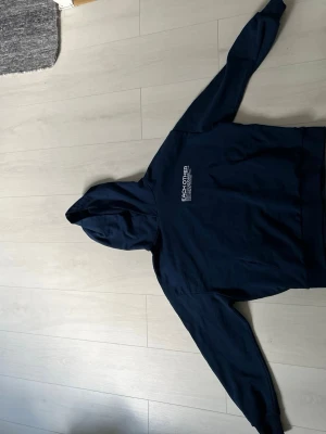 Mörkblå hoodie från Each Other - Snygg mörkblå hoodie från Each Other med vit text på bröstet. Klassisk modell med huva och känguruficka framtill. Perfekt för en avslappnad och trendig stil. 