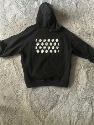 Maison Margiela svart hoodie - Säljer en svart hoodie från Maison Margiela med unik cut-out design på ryggen där loggan och siffror syns genom cirkelformade hål. Klassisk huva och ribbade muddar. Materialet är mjuk bomull som känns skön mot huden. Perfekt för dig som gillar statement-plagg.