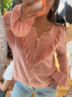 Rosa kofta med volangdetaljer - Supersöt rosa kofta med knappar framtill och volangdetaljer i V-form över bröstet. Långa ärmar med breda, ribbade muddar som ger en extra touch. Perfekt att styla med jeans för en trendig look.