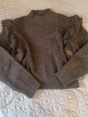 Stickad tröja med volangärmar - Säljer en brun/grå stickad tröja med höghals och snygga volangdetaljer på axlarna. Tröjan har ett fint hålmönster och ribbade muddar vid ärmslut och nederkant. Perfekt för dig som gillar unika detaljer och en trendig look.