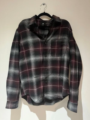 Rutig skjorta från H&M i vinrött - Snygg rutig skjorta från H&M i vinröda, svarta och vita toner. Skjortan har lång ärm, klassisk krage, bröstficka och knappar framtill. Perfekt för en avslappnad och trendig look.