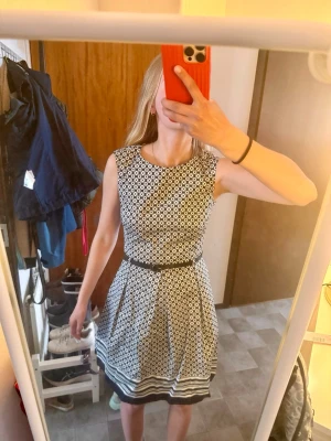 Svartvit mönstrad klänning med bälte - Snygg svartvit klänning med geometriskt mönster och markerad midja med smalt bälte. Klänningen är ärmlös, har rund halsringning och slutar ovanför knäna. Perfekt för dig som gillar klassisk stil med en twist.