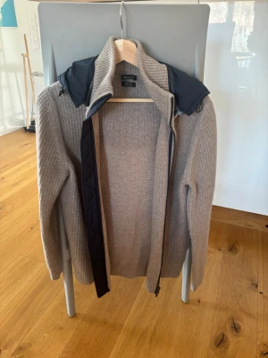 Beige stickad cardigan Massimo dutti  - Säljer en beige stickad tröja från Massimo Dutti med svart huva och dragkedja framtill. Tröjan har lång ärm och ribbad struktur, perfekt för dig som gillar en stilren och avslappnad look.