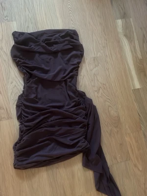 Mörklila omlottklänning med drapering - Snygg mörklila kortklänning med omlott-detalj och draperad design. Klänningen är axelbandslös och har en lång tygdel som hänger ner på sidan för en extra cool look. Perfekt för dig som vill sticka ut med en trendig och elegant stil.