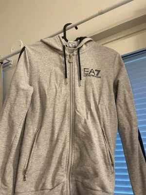 Grå hoodie från EA7 Emporio Armani - Snygg grå hoodie med dragkedja från EA7 Emporio Armani. Hoodien har svarta detaljer på axlarna, två fickor med dragkedja (ena sönder, se bild). 