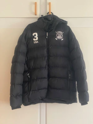 Svart pufferjacka från Vinson Polo Club - Svart pufferjacka med huva från Vinson Polo Club. Jackan har vita tryck med texten '3 USA' och klubbens logga på bröstet. Dragkedja framtill och två sidofickor med dragkedja. Perfekt för kalla vinterdagar.