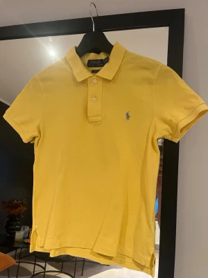 Gul pikétröja från Polo Ralph Lauren - Snygg gul pikétröja från Polo Ralph Lauren med klassisk krage, korta ärmar och blå broderad logga på bröstet. Tröjan är i bomull och har knappar framtill. Perfekt för en avslappnad och stilren look.