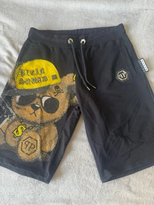 Philipp Plein shorts med björntryck - Svarta shorts från Philipp Plein med stort färgglatt björntryck framtill, dekorerade med glittriga stenar. Shortsen har snörning i midjan och en logotypdetalj på ena benet. Materialet känns mjukt och bekvämt, perfekt för en avslappnad stil. Storlek L men skulle säga passar S/M. Köpta på NK