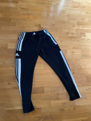 Svarta Adidas träningsbyxor AEROREADY - Svarta träningsbyxor från Adidas med vita ränder längs sidorna och AEROREADY-tryck vid midjan. Byxorna har elastisk midja och smal passform, perfekta för sport och träning. Tillverkade i ett lätt och ventilerande material. 3 st!!! 100kr st