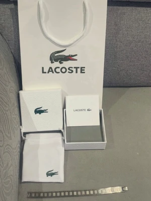 Silverfärgat armband från Lacoste - Stilrent armband från Lacoste i silverfärgad metall med klassisk krokodil-logga ingraverad på spännet. Armbandet har en modern, fyrkantig länkdesign och levereras med originalförpackning och påse.