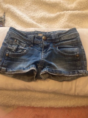 Blå jeansshorts med dragkedjor - Snygga blå jeansshorts med klassisk femficksdesign, uppvikta benslut och coola dragkedjor på bakfickorna. Shortsen har bälteshällor och knappstängning framtill. Perfekta för varma dagar och en avslappnad stil.