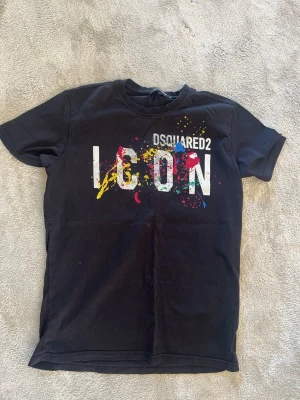 Svart Dsquared2 ICON t-shirt - Cool svart t-shirt från Dsquared2 med stort vitt 'ICON'-tryck och färgglada färgstänk på bröstet. Klassisk rund hals och korta ärmar. Perfekt för dig som vill sticka ut med en trendig streetstyle-look.