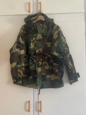 Klassisk camo vindjacka med huva - Säljer en vindjacka i klassiskt militärinspirerat camomönster i grönt, brunt och svart. Jackan har huva, dragsko nedtill och justerbara ärmslut. Perfekt för dig som gillar streetwear eller outdoor-vibe. Materialet är tunt och vindtätt, perfekt för blåsiga dagar.