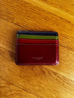 Korthållare - Säljer en stilrent korthållare i älta läder med flera fack. Framsidan har röd, grön och mörkblå färgblock och baksidan är helröd med en liten dragkedjeficka. Perfekt för kort och mynt, med vita kontrastsömmar för en snygg touch.