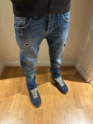 Blå slitna jeans med hål från dondup   - Säljer ett par blå jeans från dondup  med avslappnad passform och slitna detaljer, inklusive hål på framsidan och bakfickor med unik design. Jeansen har klassisk femficksmodell och är gjorda i denim. 
