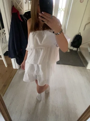 Vit klänning - Säljer en vit axelbandslös kortklänning med volangdetaljer. Klänningen har en luftig och flowy passform, perfekt för konfa/student eller skolavslutning. Materialet är lite mer åt det lyxiga hållet och klänningen har en enkel, stilren design utan mönster. One shoulder strl xs använd 2 gånger och egensydd av uf företag 💞