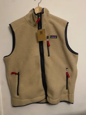 Patagonia fleece vest - Beige Patagonia fleece vest – retro outdoor style  Beige / khaki fleece vest from Patagonia made of soft and warm teddy fleece material. Features a full zip, high collar, and multiple zippered pockets, including a chest pocket. Contrast navy details and red zipper pulls give it a classic retro outdoor look.  Perfect as a layering piece or worn over a hoodie or sweater. Suitable for everyday wear, hiking, and outdoor activities.   Color: Beige / khaki Brand: Patagonia Fit: Classic Condition: New