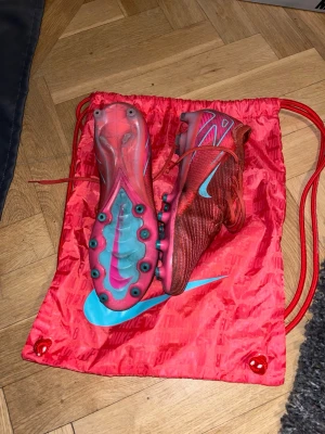 Nike Mercurial röda fotbollsskor AG - Säljer ett par Nike Mercurial fotbollsskor i rött med turkosa detaljer och logga. Låda och påse kommer med. Använda ca 5 matcher endast, inga skador.