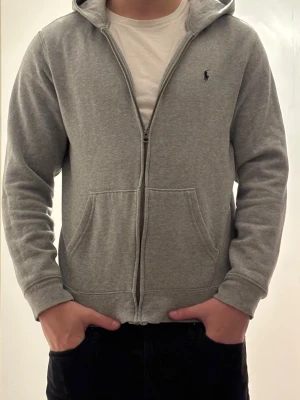 Grå hoodie med dragkedja från Polo Ralph Lauren - Snygg grå hoodie från Polo Ralph Lauren med dragkedja framtill och klassisk huva. Storlek barn XL men sitter som S 