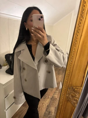 Kort beige trenchcoat - Snygg kort trenchcoat i beige med dubbelknäppning och stora bruna knappar. Jackan har breda slag och justerbara ärmslut med knappar. Perfekt för att ge en stilren look och passar till många outfits.