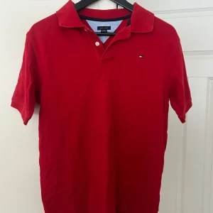 Röd pikétröja från Tommy Hilfiger - Snygg röd pikétröja från Tommy Hilfiger med korta ärmar och klassisk krage. Perfekt för en stilren och avslappnad look. Den har en liten logotyp på bröstet och är tillverkad i ett bekvämt material.