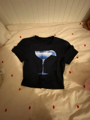 Marinblå croppad t-shirt med glastryck - Cool marinblå croppad t-shirt med ett blått tryck av ett cocktailglas på bröstet. Perfekt för dig som vill sticka ut med en unik och trendig look. T-shirten har korta ärmar och rund halsringning.